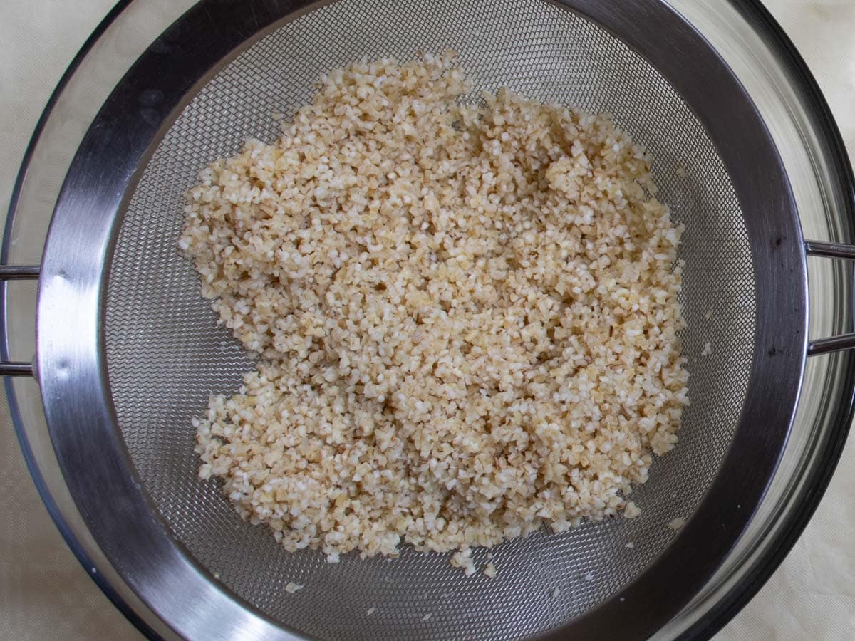 Soaked bulgur wheat being drained in metal sieve suspended over glass bowl.