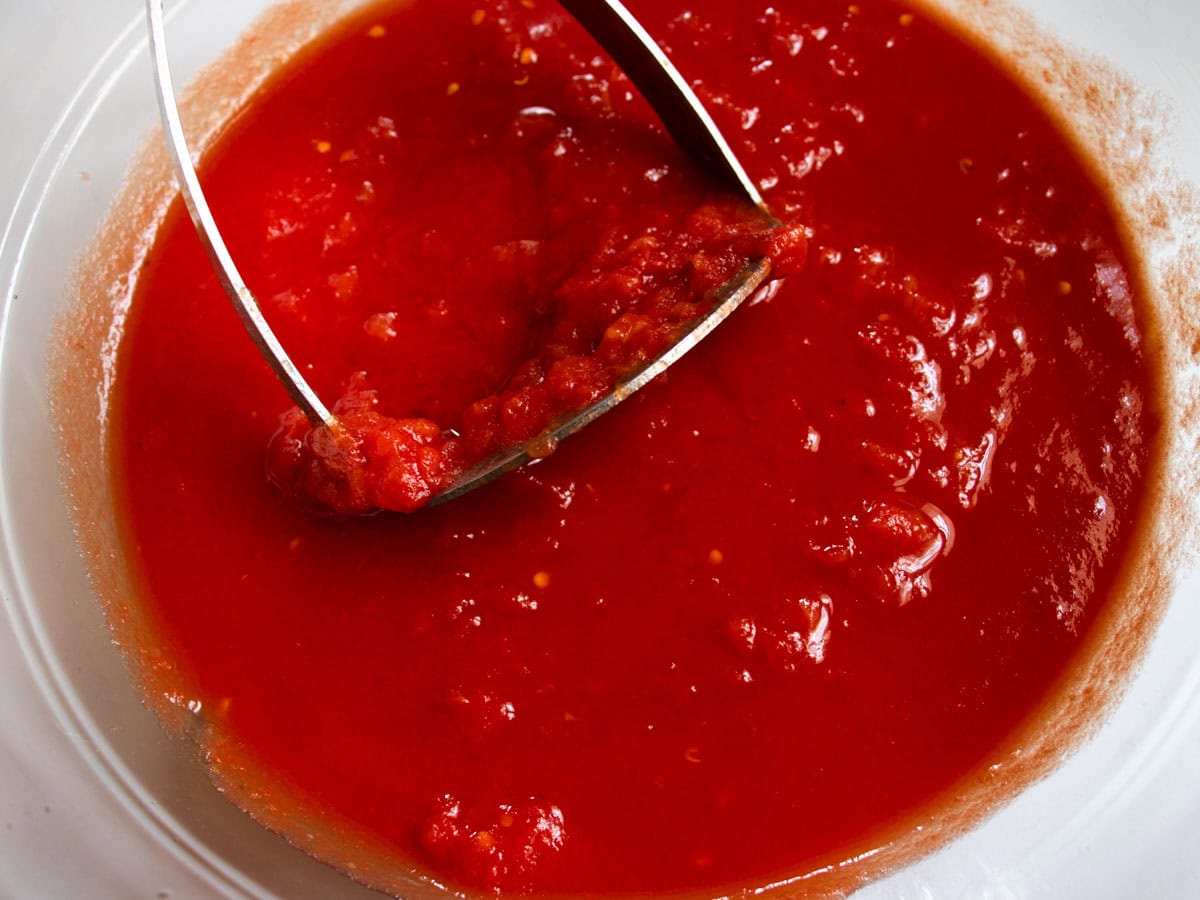Canned tomatoes in glass bowl being crushed by potato masher.
