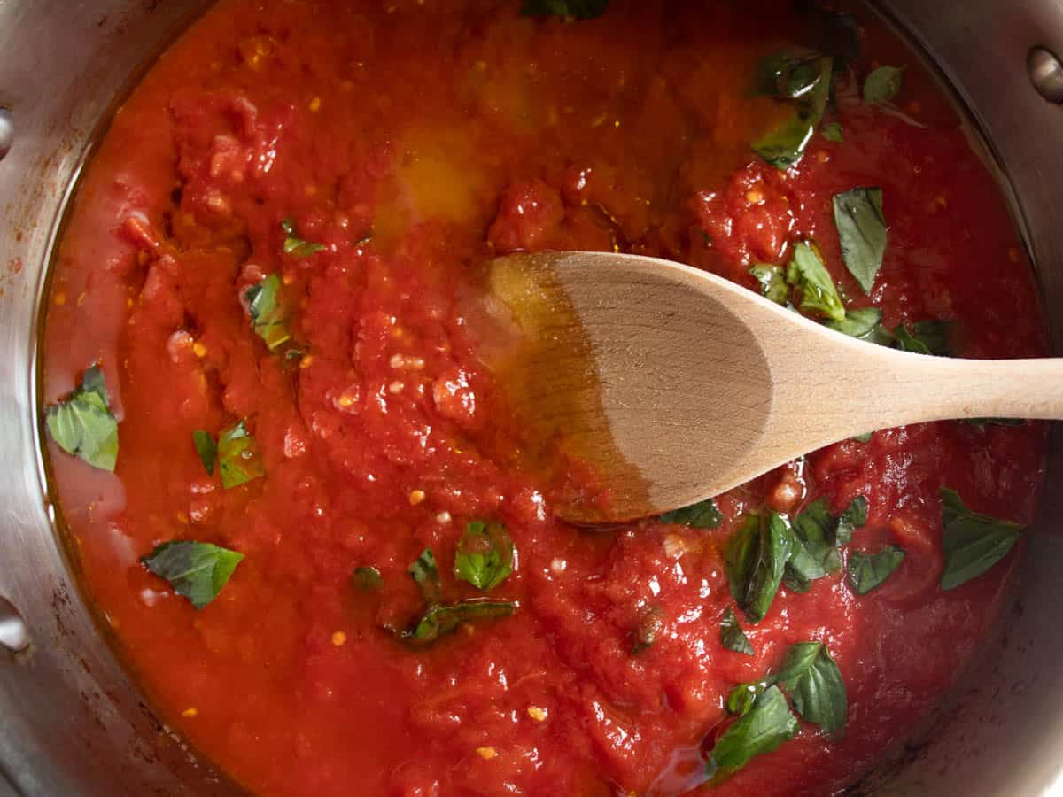 Crushed tomatoes, olive oil and torn basil leaves being stirred in stainless steel sauce with wooden spoon.