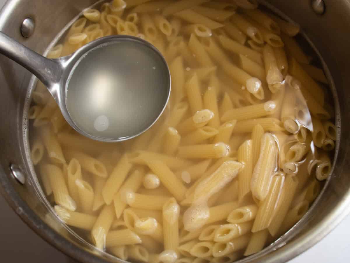 Ladle filled with pasta water being held over stainless steel saucepan full of cooked pasta in water.
