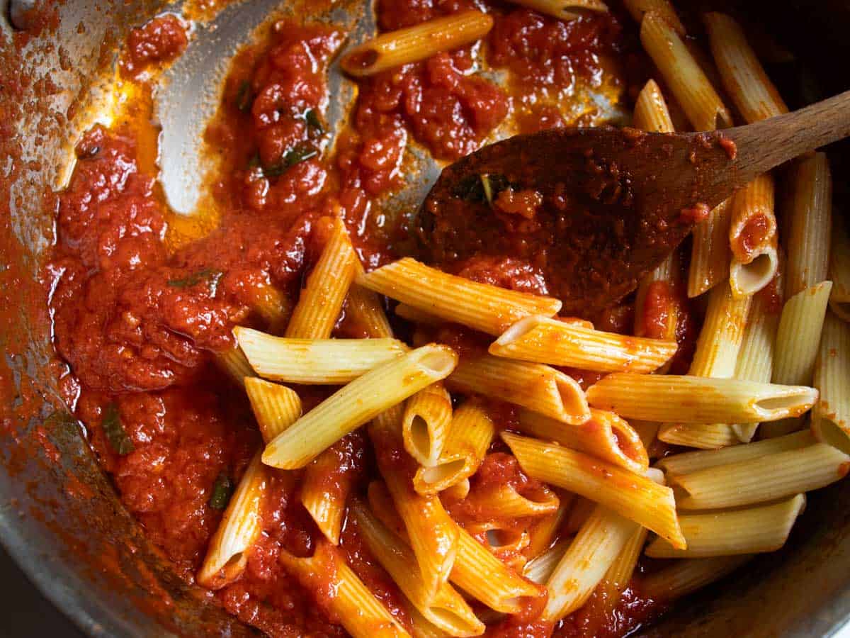 Penne pasta being stirred into Napoli tomato sauce with wooden spoon in stainless steel saucepan.