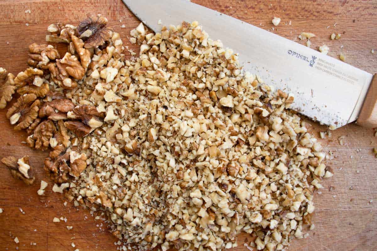 Walnuts halves and walnut pieces on wooden board next to chef's knife.