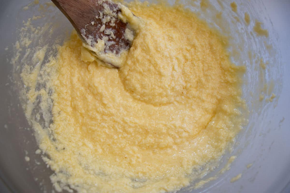 Creamed butter with eggs mixture being stirred with wooden spoon in glass mixing bowl.