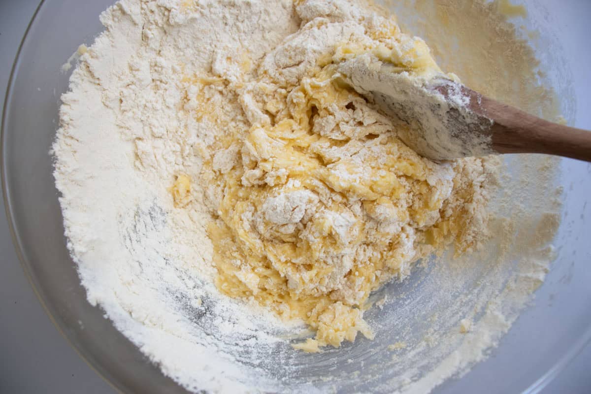Flour being stirred into sugar, butter, egg mixture by wooden spoon in glass mixing bowl.