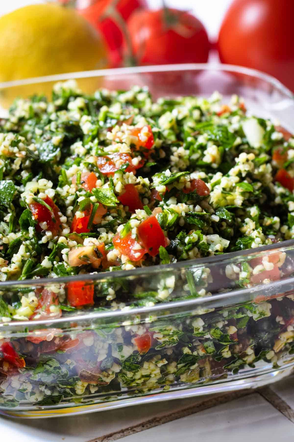 Lebanese tabbouleh in glass dish on tiled surface, lemon and tomatoes in background.