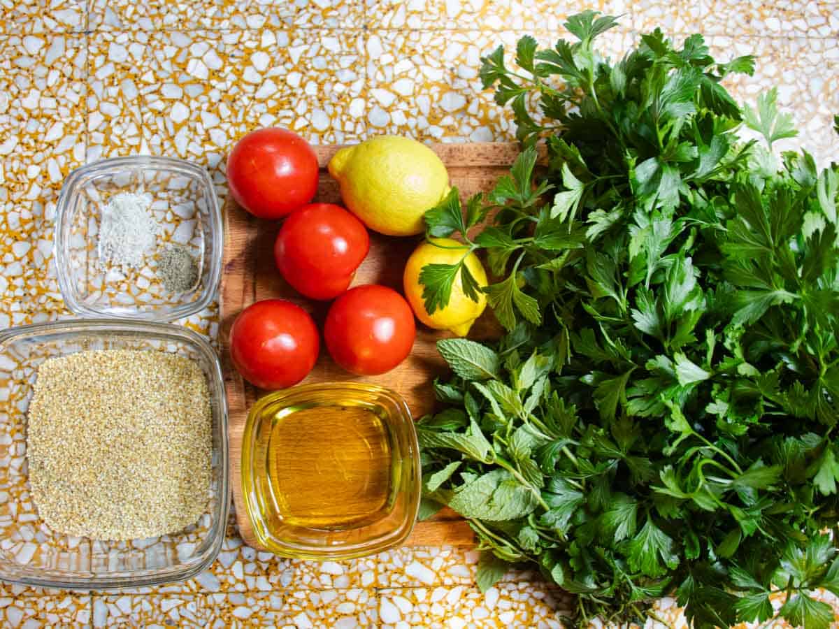 Tomatoes, lemons and bunches of mint and parsley, together with oil in glass dish laid out on wooden board, next to bulgur wheat in glass dish and salt and pepper.