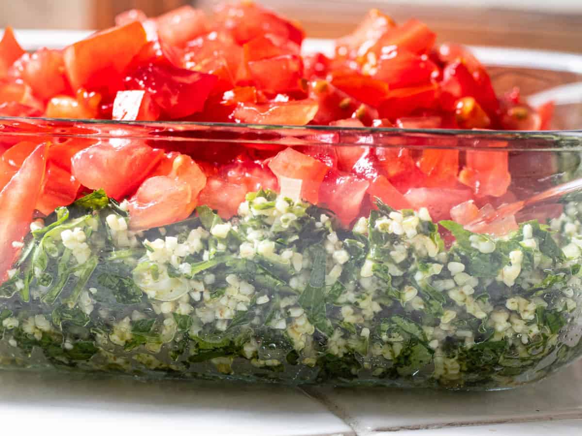 Diced chopped tomatoes on bed of parsley, mint and bulgur tabbouleh mix in glass dish on tiled surface.
