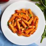 Penne Napoli pasta in white dish next to sprig of basil and blue cloth.