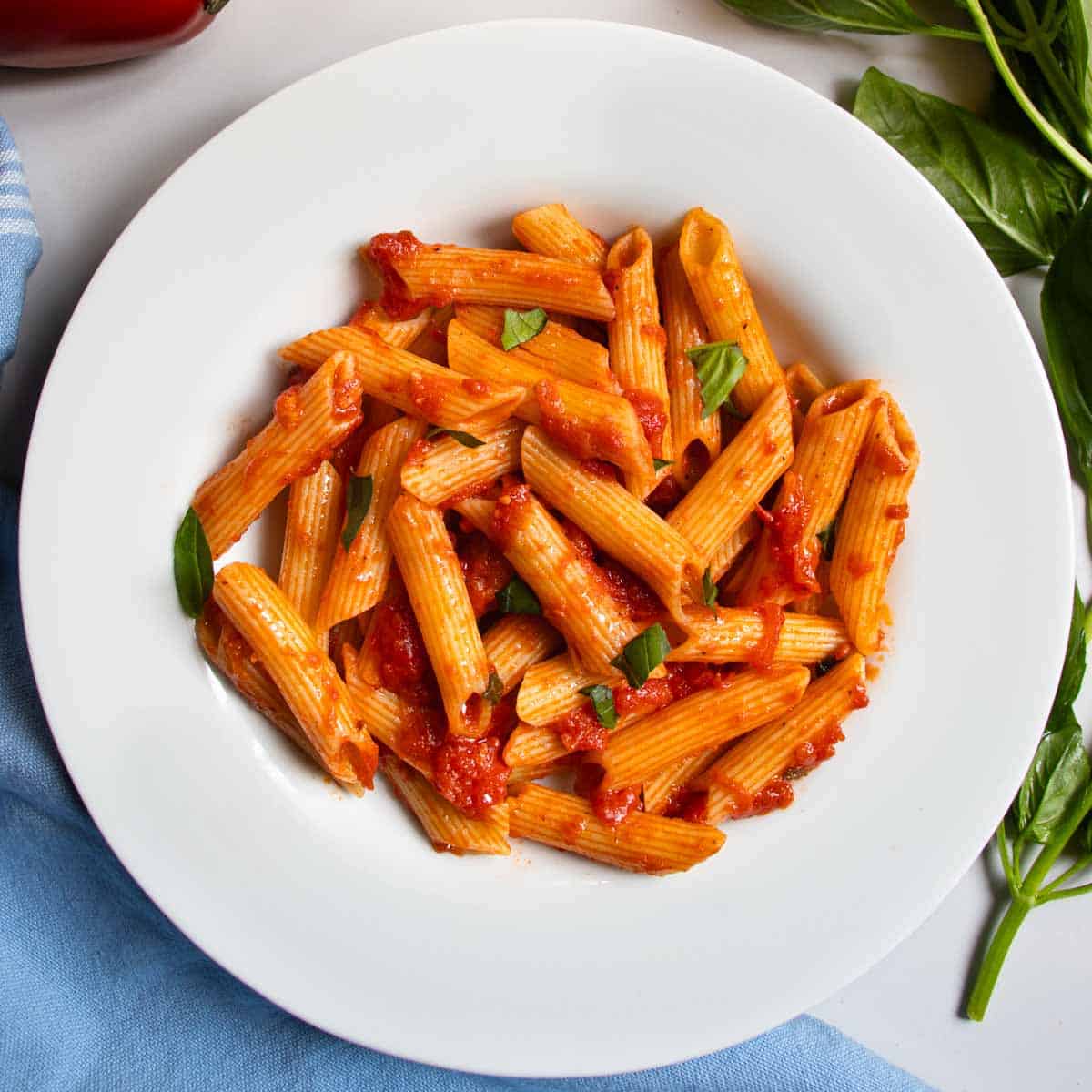 Penne Napoli pasta in white dish next to sprig of basil and blue cloth.