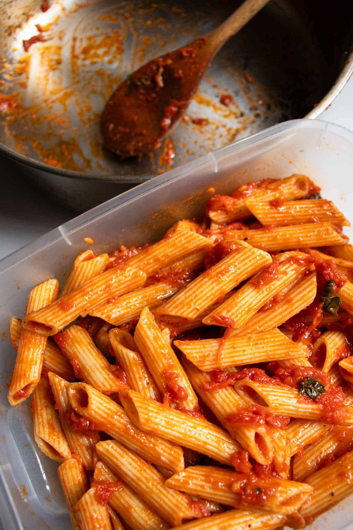 Plastic storage container filled with Penne Napoli pasta placed in front of empty stainless steel saucepan with remains of pasta sauce.