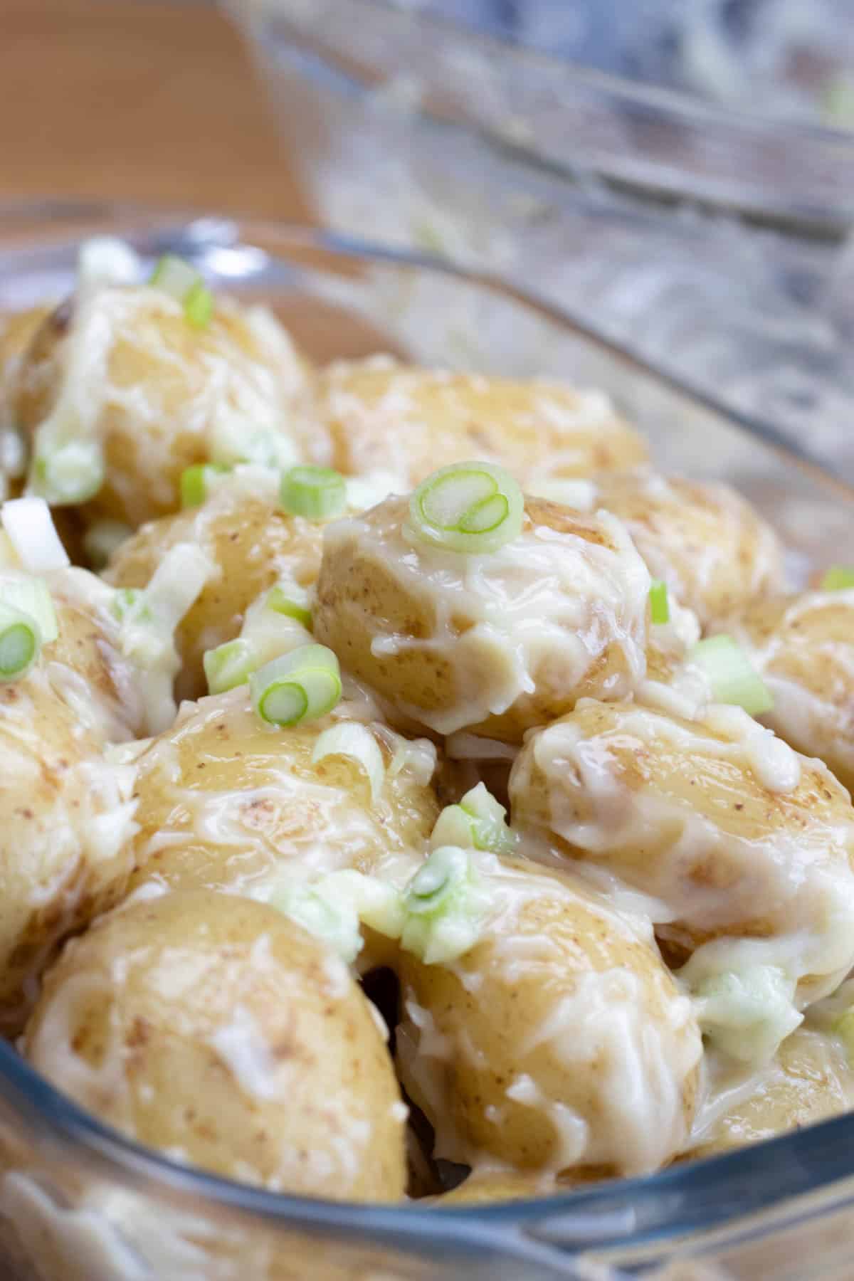 Potato salad with mayonnaise and spring onions in glass dish, empty mixing bowl with traces of mayonnaise in background.