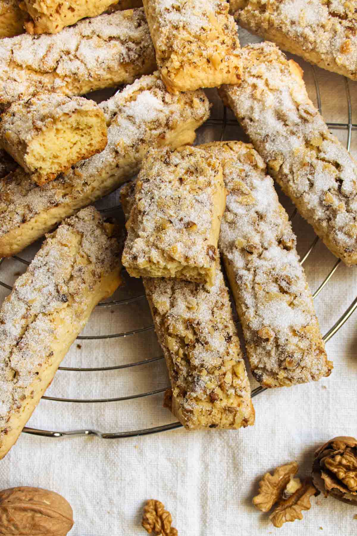Sugar-coated walnut cookies on wire tray, one broken in half, cracked walnut kernels in the foreground.