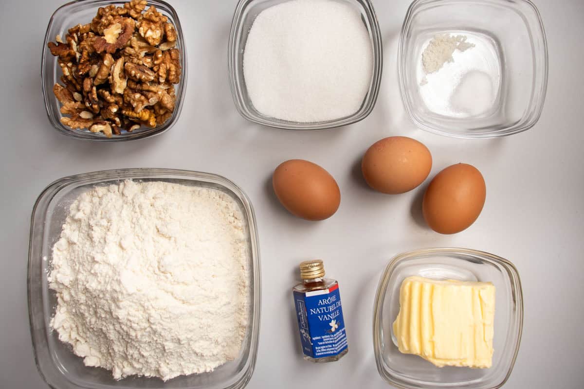 Walnuts, flour, sugar, salt, butter and bicarbonate of soda all in glass dishes, bottle of vanilla essence and eggs laid out.