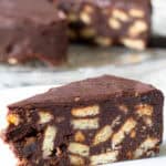 Slice of chocolate biscuit cake in foreground, whole cake from which slices have been removed showing in background.