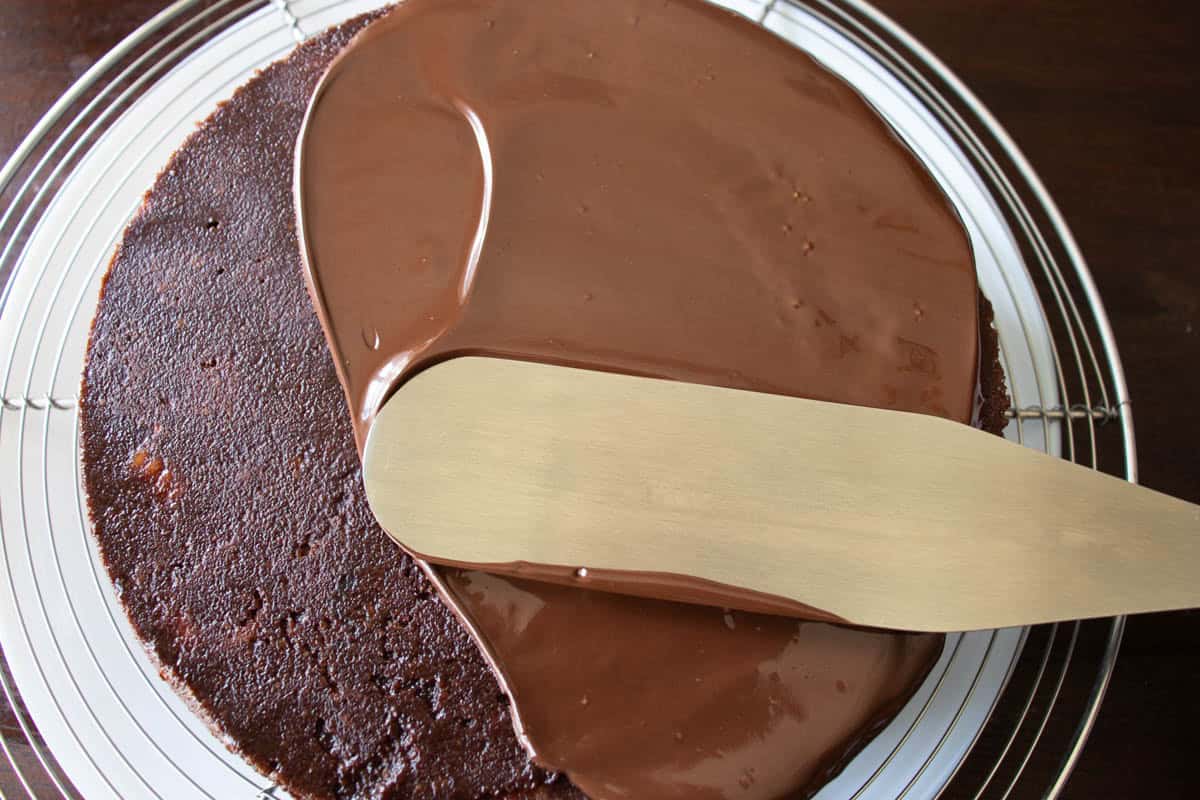 Spatula spreading melted chocolate onto chocolate biscuit cake which is on round wire tray.