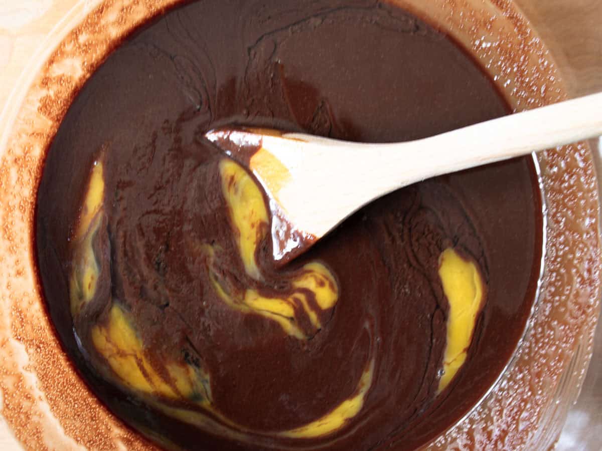 Egg being stirred by wooden spoon into chocolate mixture in glass mixing bowl.