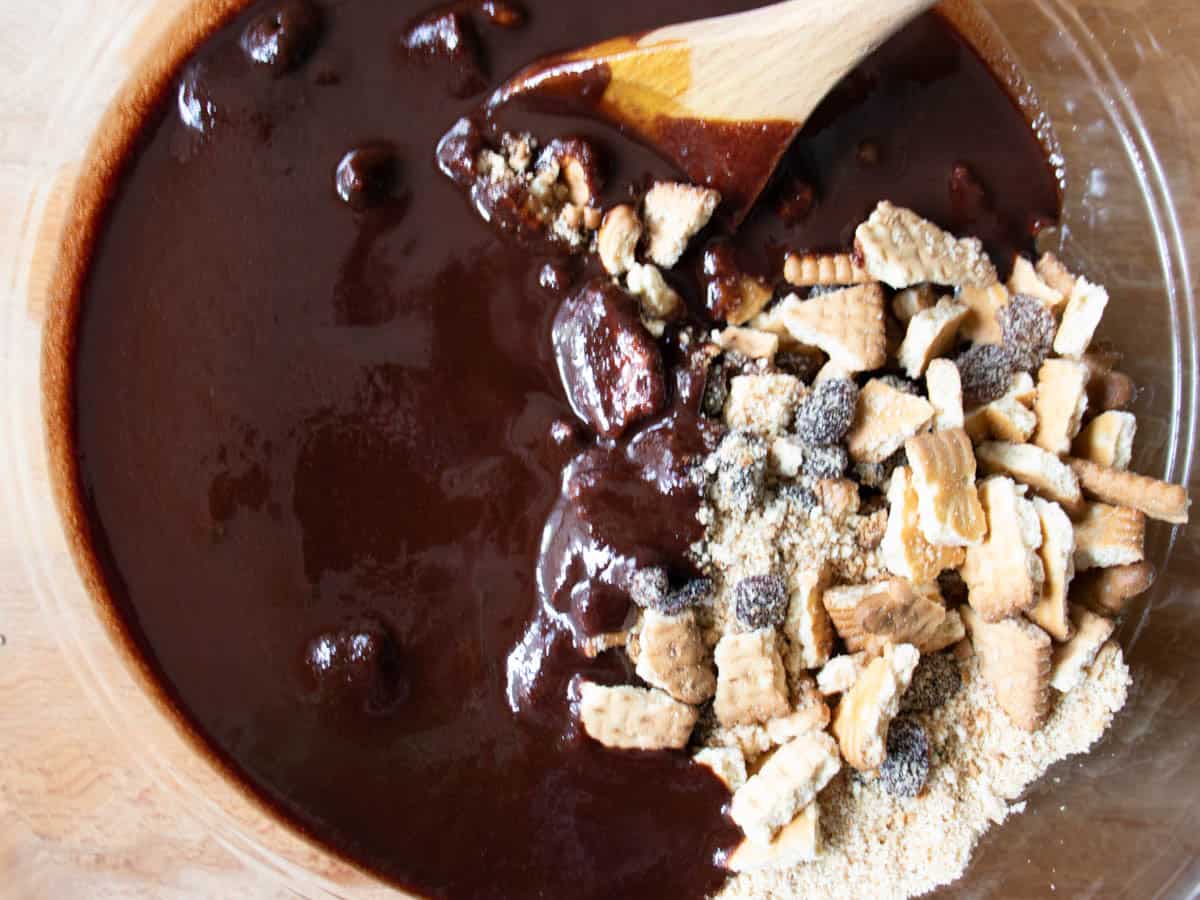 Broken biscuits and raisins being stirred by wooden spoon into chocolate mixture in glass mixing bowl.