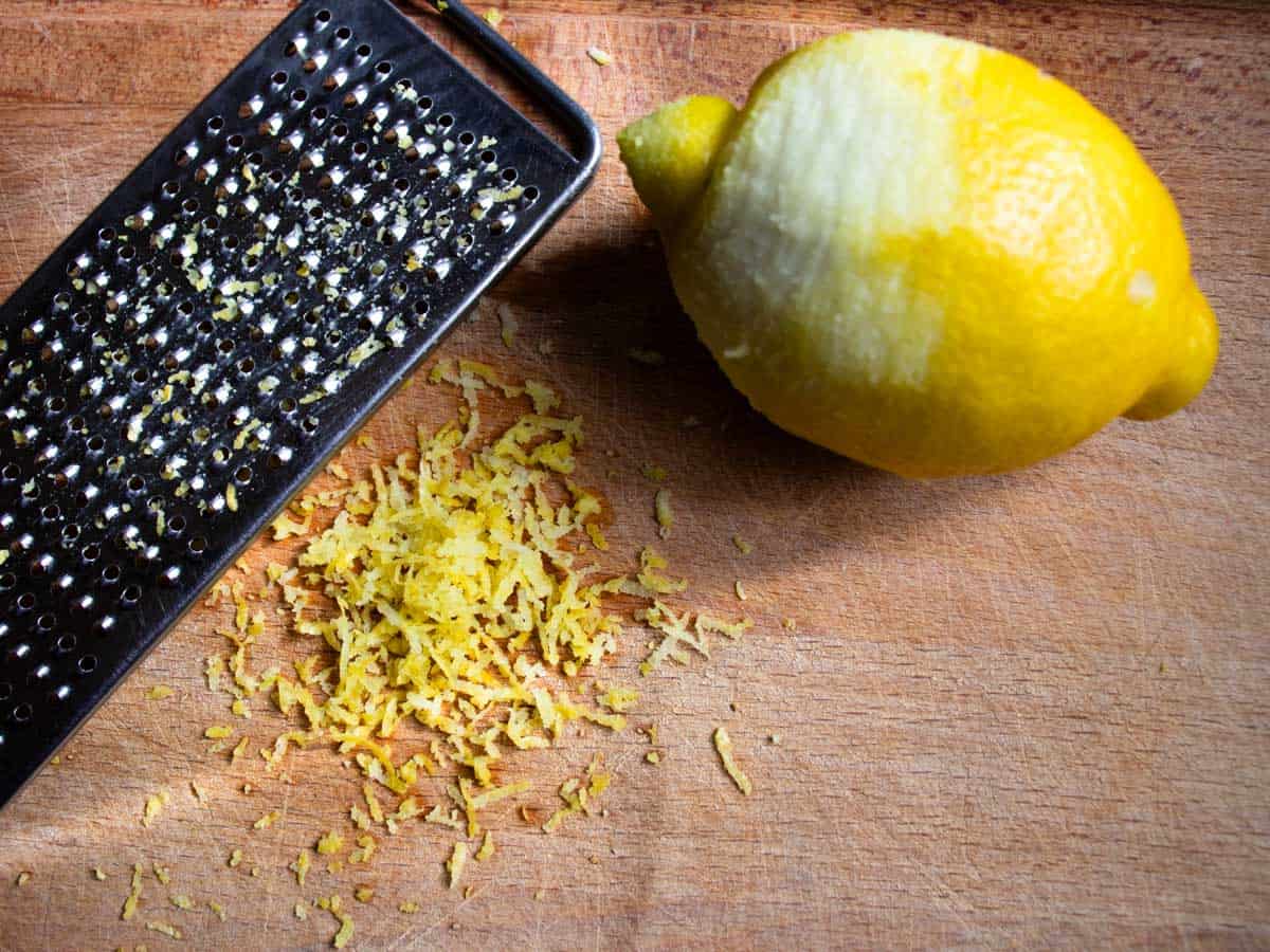 Hand-held grater next to finely grated lemon zest and half grated lemon on a wooden board.