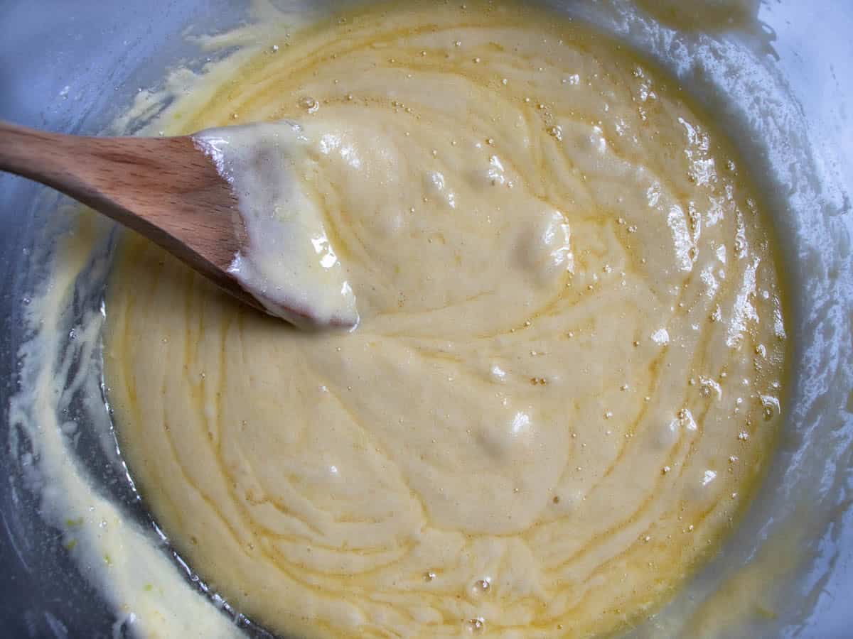 Wooden spoon being stirred through French lemon madeleines batter in glass mixing bowl.