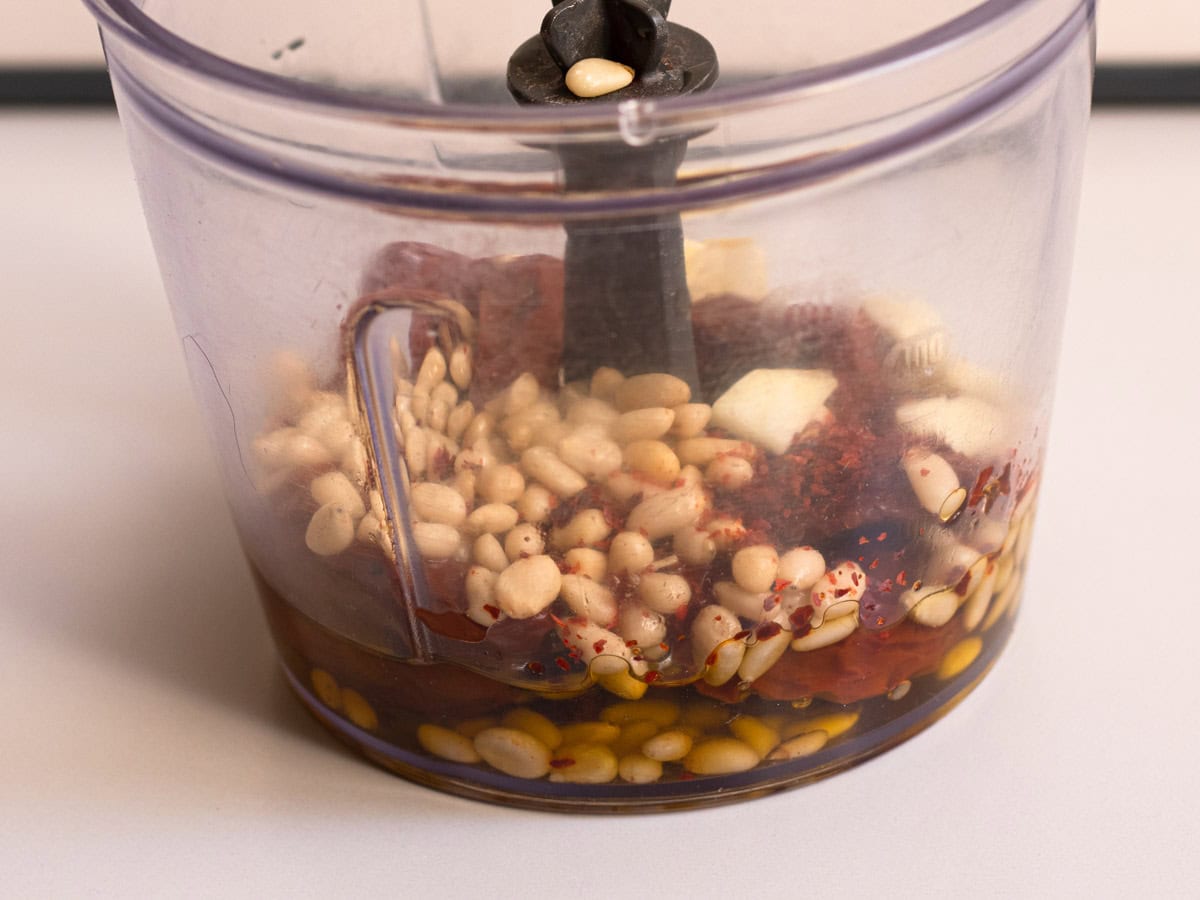 Side view of plastic blender container with pine nuts, sun-dried tomatoes, olive oil, chilli flakes and chopped garlic before blending.