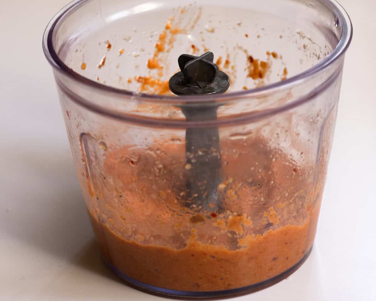 Side view of plastic blender container with pine nuts, sun-dried tomatoes, olive oil, chilli flakes and chopped garlic after blending.
