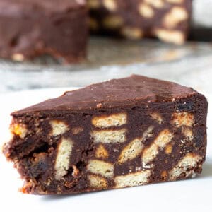 Slice of chocolate biscuit cake in foreground, whole cake from which slices have been removed showing in background.