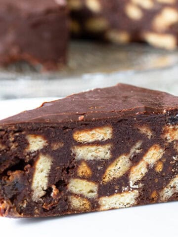 Slice of chocolate biscuit cake in foreground, whole cake from which slices have been removed showing in background.