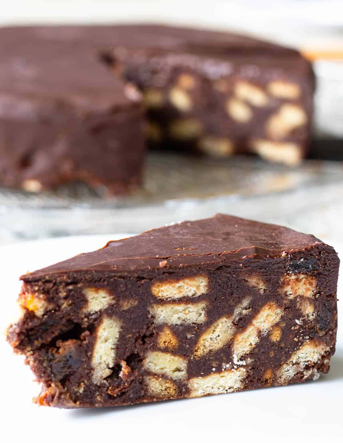 Slice of chocolate biscuit cake in foreground, whole cake from which slices have been removed showing in background.