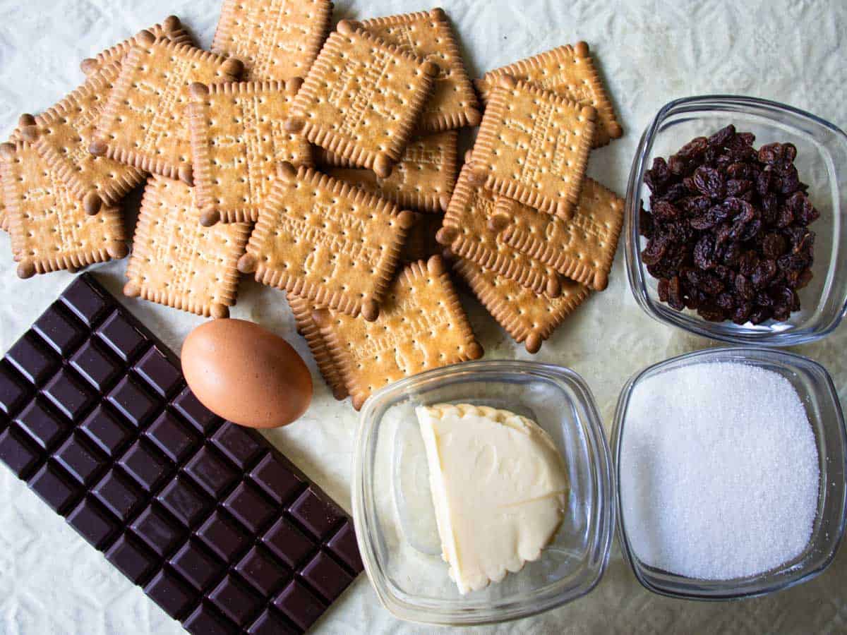Ingredients for no-bake chocolate biscuit cake laid out, petit beurre biscuits, chocolate, egg, butter, raisins and sugar.