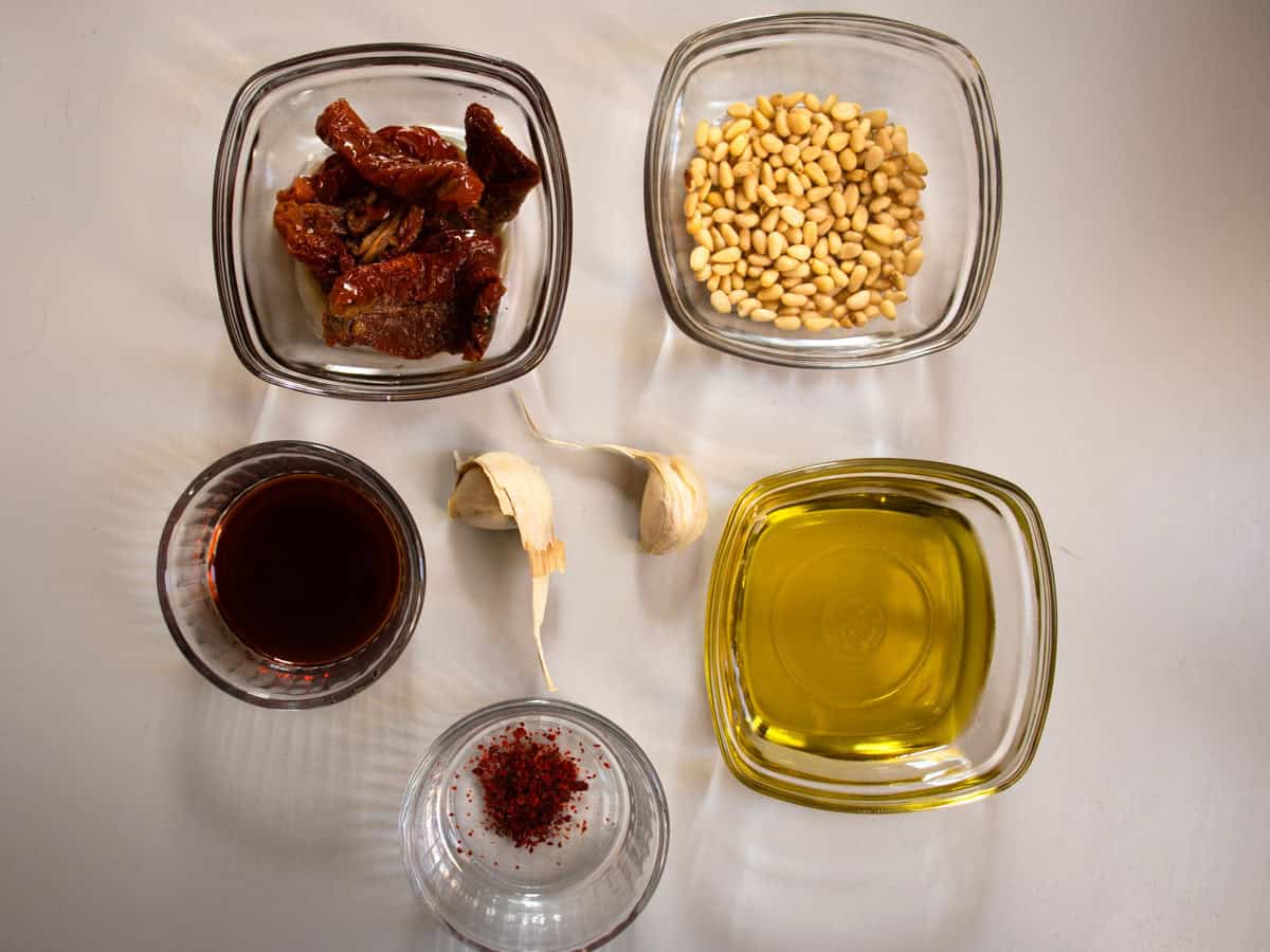 Ingredients for red pesto sauce laid out in glass dishes, sun-dried tomatoes, pine nuts, vinegar, chilli flakes, olive oil and garlic.