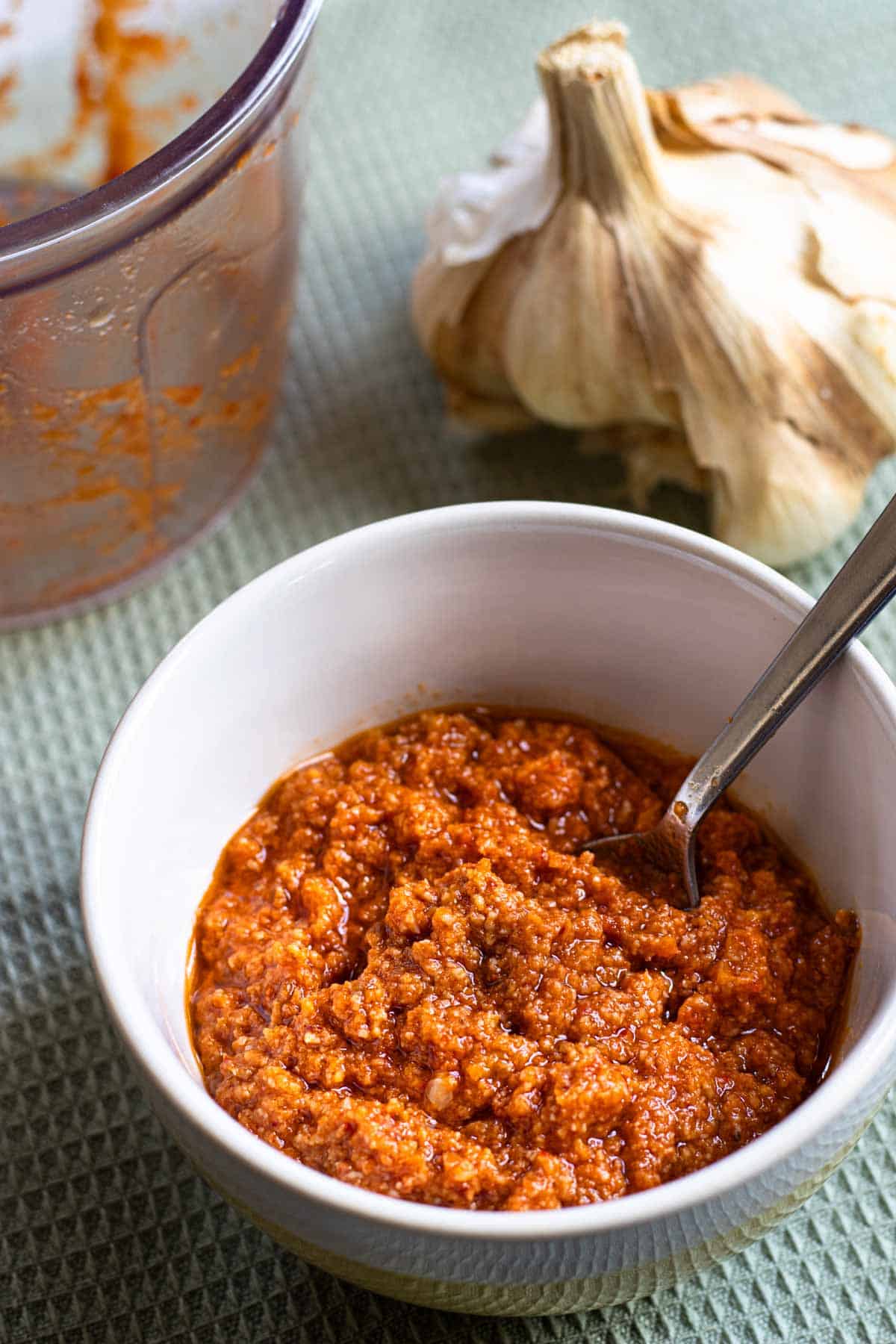 Red pesto sauce in small bowl, with spoon in it, garlic bulb and used plastic blender container in background.